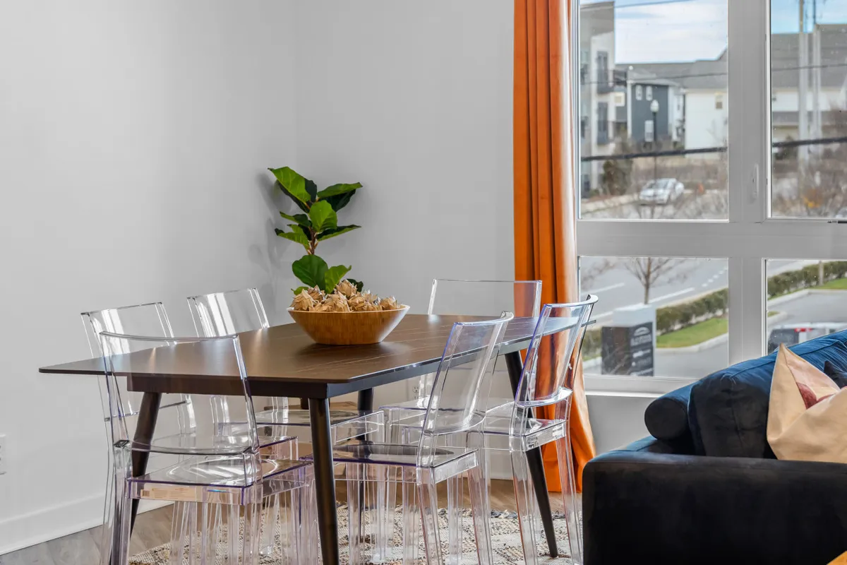 A modern dining area with a sleek table, transparent chairs, a decorative bowl centerpiece, an indoor plant, and a large window draped with airy curtains. The space is bathed in light, perfect for meals, games, or planning outings.