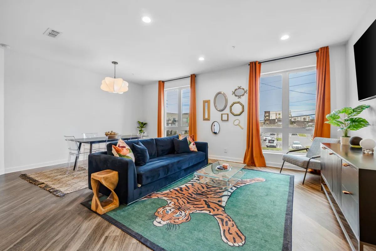A playful yet stylish living area with a blue sofa, eye-catching tiger rug, glass coffee table, orange curtains, and decorative mirrors. The setup is ideal for relaxing, socializing, and movie nights, with artistic touches that add personality.