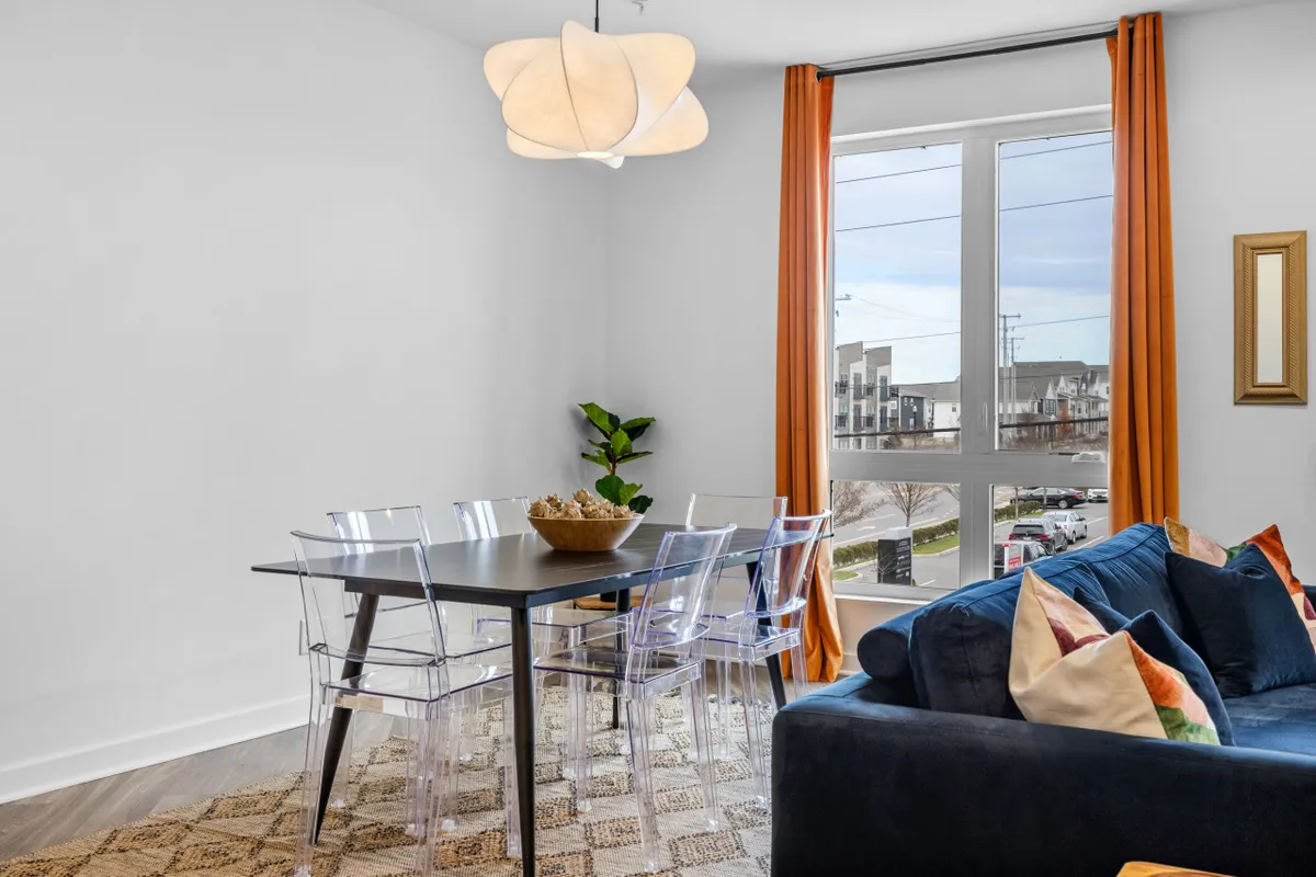 A welcoming dining area with a stylish chandelier, a sunlit window, a comfortable sofa with cushions, and a dining table ready for shared meals or conversations. The scene encourages socializing, whether over dinner or coffee, and hints at the vibrant city atmosphere just outside.