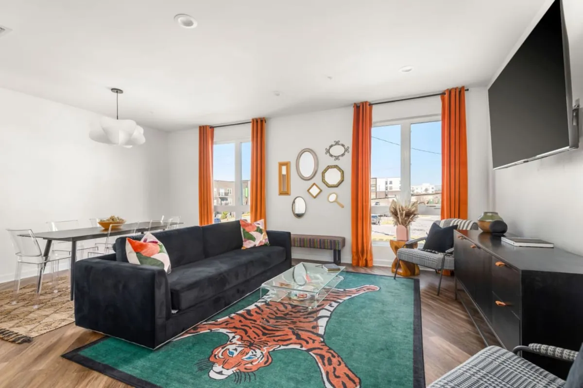 The photo showcases a lively living space featuring a sleek black couch, a playful tiger rug, bold orange curtains, a modern glass coffee table, and a wall-mounted TV. The artwork and decor add a vibrant, energetic feel, suggesting a perfect spot for groups to gather before heading out or after a night on the town.