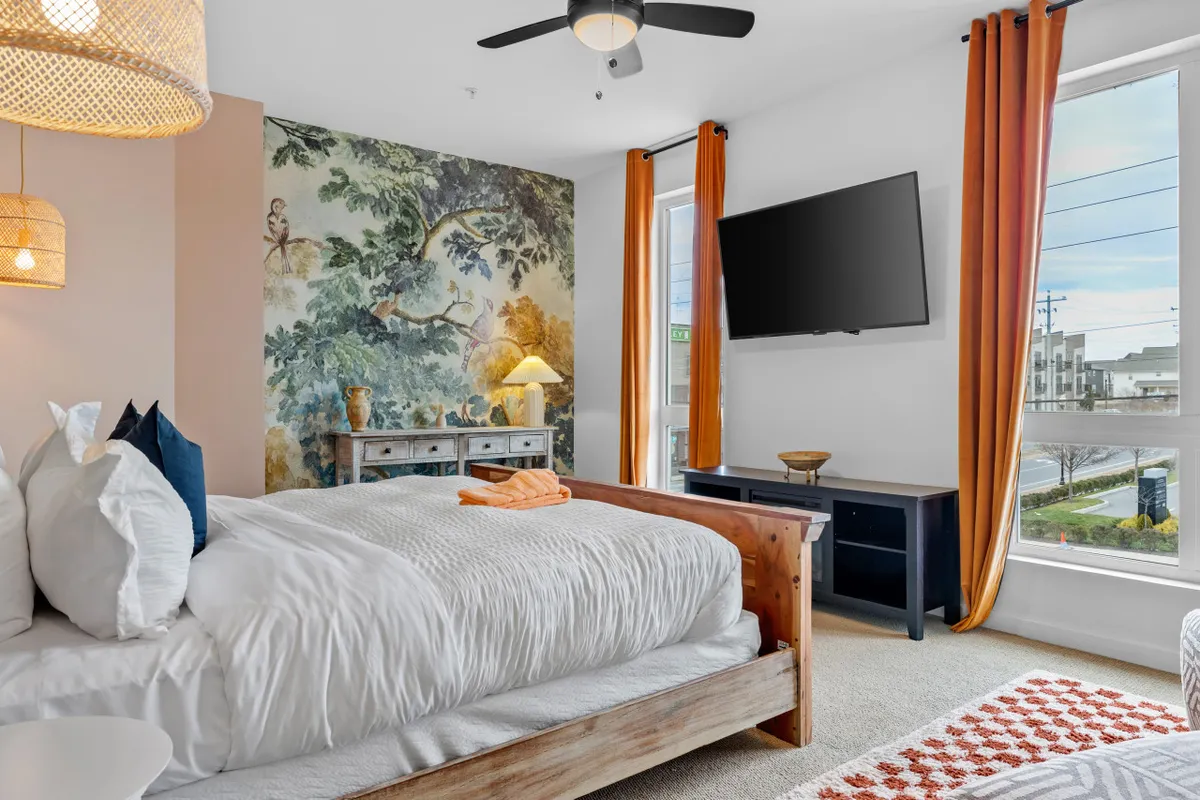 A cozy bedroom featuring a king-sized bed with fluffy pillows, a large mural on the wall, orange curtains framing the window, and a TV mounted on the wall. Sunlight bathes the space, highlighting the calm and inviting atmosphere. The room is designed for relaxation, with a reading nook and the convenience of in-bed entertainment.