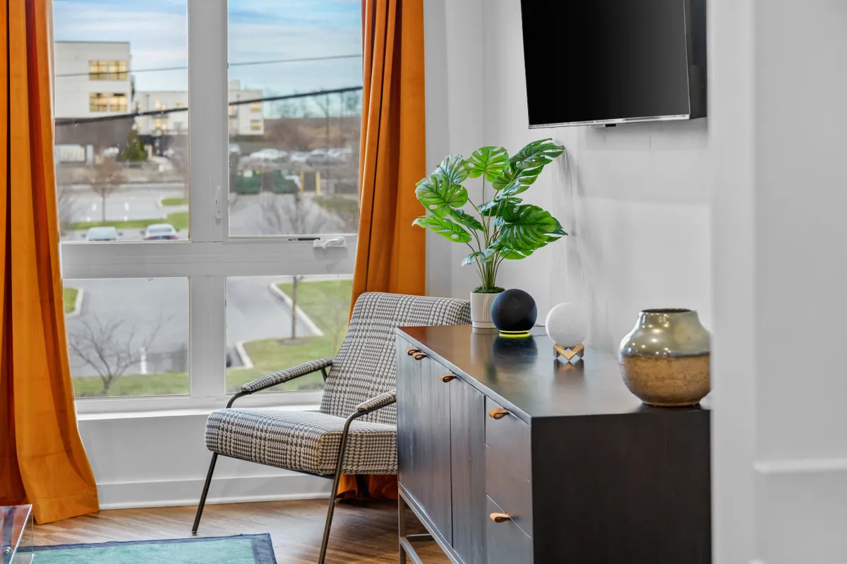 A cozy, modern living area featuring a window with vibrant orange curtains that lets in natural light. There is a stylish chair positioned for reading or relaxing, a decorative plant providing a touch of greenery, a sleek wooden sideboard, and a TV set up for entertainment. The scene is inviting, suggesting a peaceful spot to unwind after exploring the city.