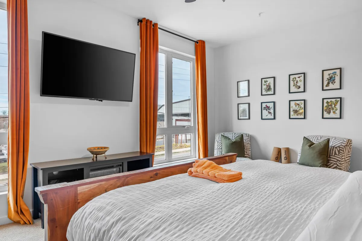A stylish bedroom with a king-sized bed dressed in textured bedding, complemented by orange curtains and a large TV on the wall. Framed floral art above the bed adds a touch of elegance, while natural light streams in, creating a restful and cozy vibe ideal for winding down.