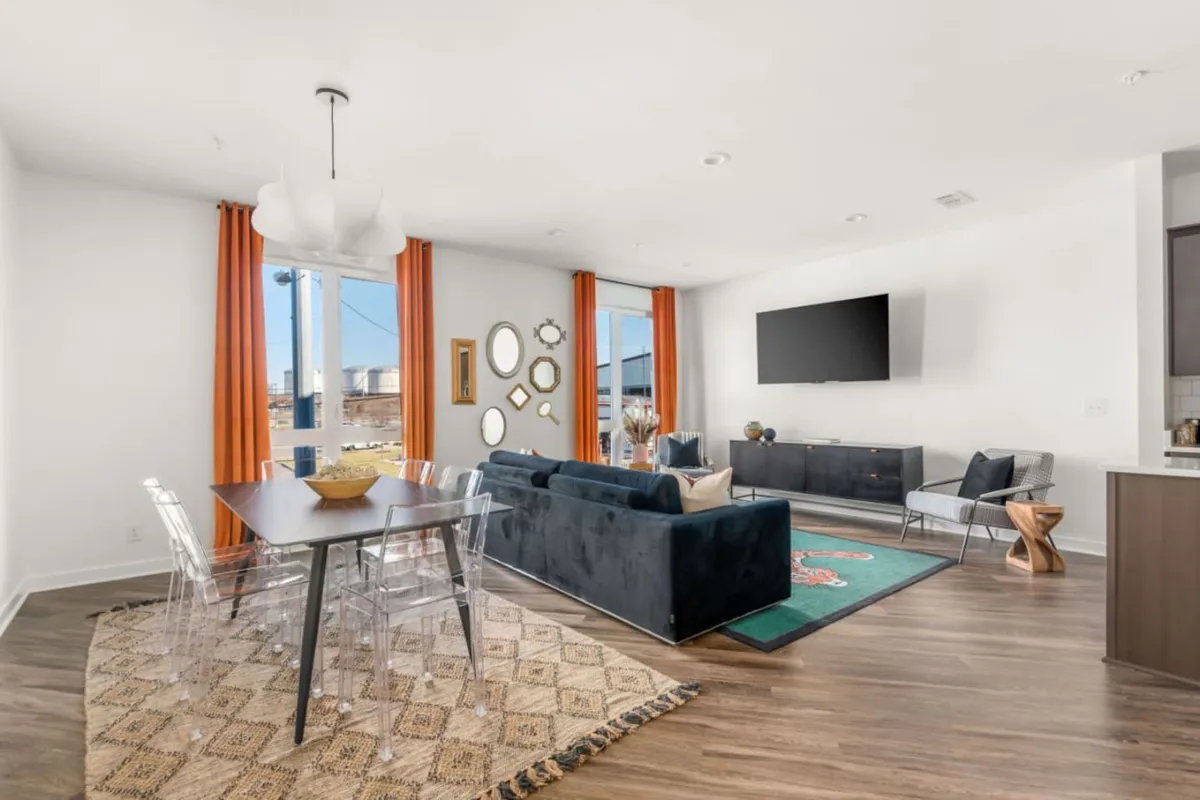 A spacious, open-plan living and dining area featuring a plush blue sofa, a colorful rug, a large flat-screen TV, a dining table with transparent chairs for six, and vibrant orange curtains. The room is filled with natural light from tall windows, creating an inviting and lively atmosphere.