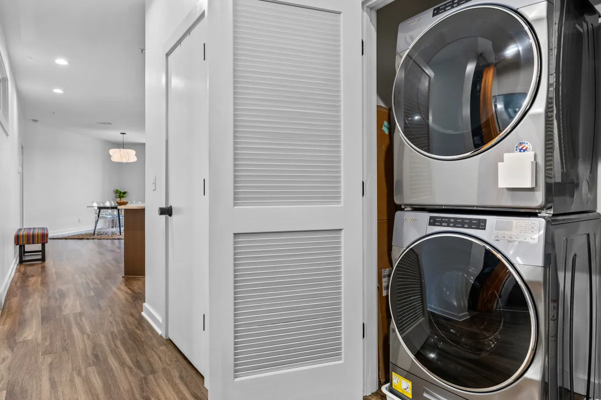 A practical laundry area with a stacked washer and dryer set, positioned near an open doorway that leads to the main living space. A decorative chair in the background adds a cozy touch, and the setup feels organized and accessible for daily chores.