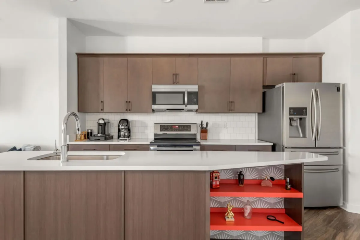 This image features a bright and stylish kitchen with a central island, stainless steel appliances, and attractive wood cabinetry. The colorful shelf adds a touch of playfulness, while the open layout hints at easy entertaining and conversation. The space is set up for both cooking and social gatherings, with clear sightlines to adjacent living areas.