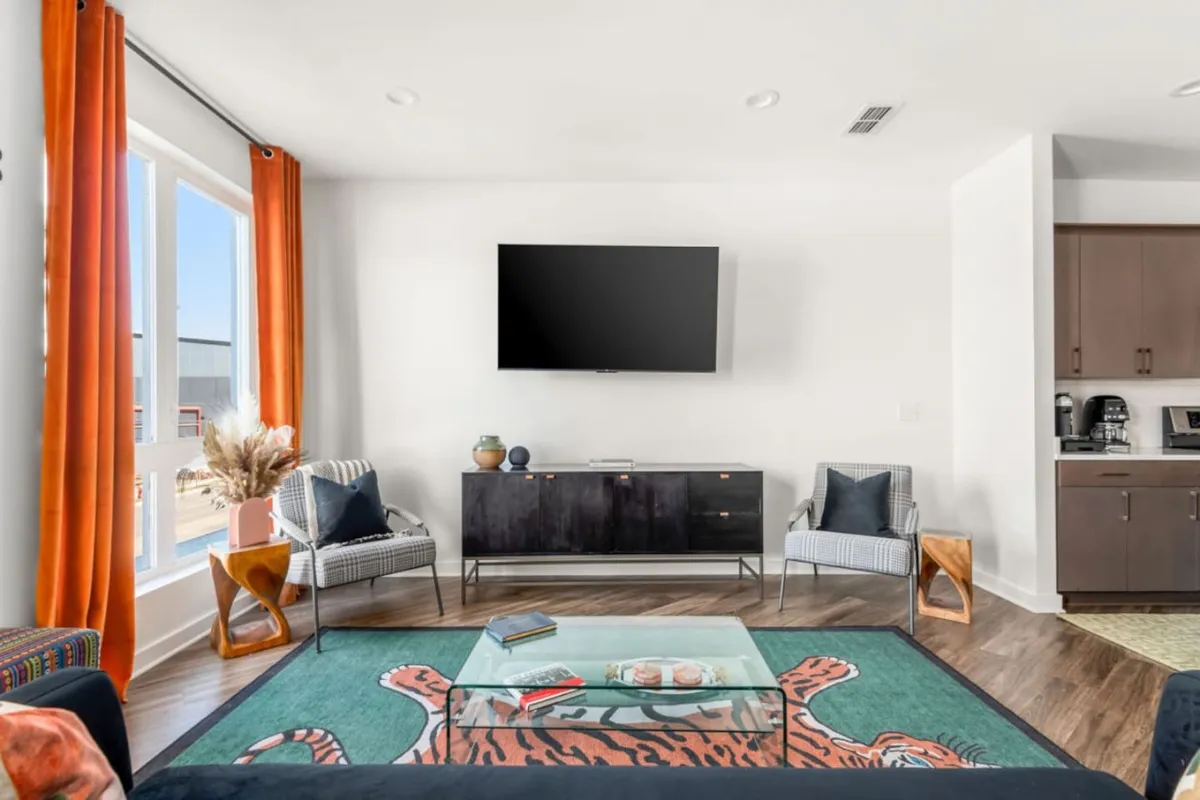 The image showcases a modern, sunlit living room with large windows framed by vibrant orange curtains, a plush seating area, a stylish glass coffee table, a bold tiger-patterned rug, and a sleek TV mounted on the wall. The overall vibe is colorful, inviting, and perfect for relaxing or gathering with friends and family.