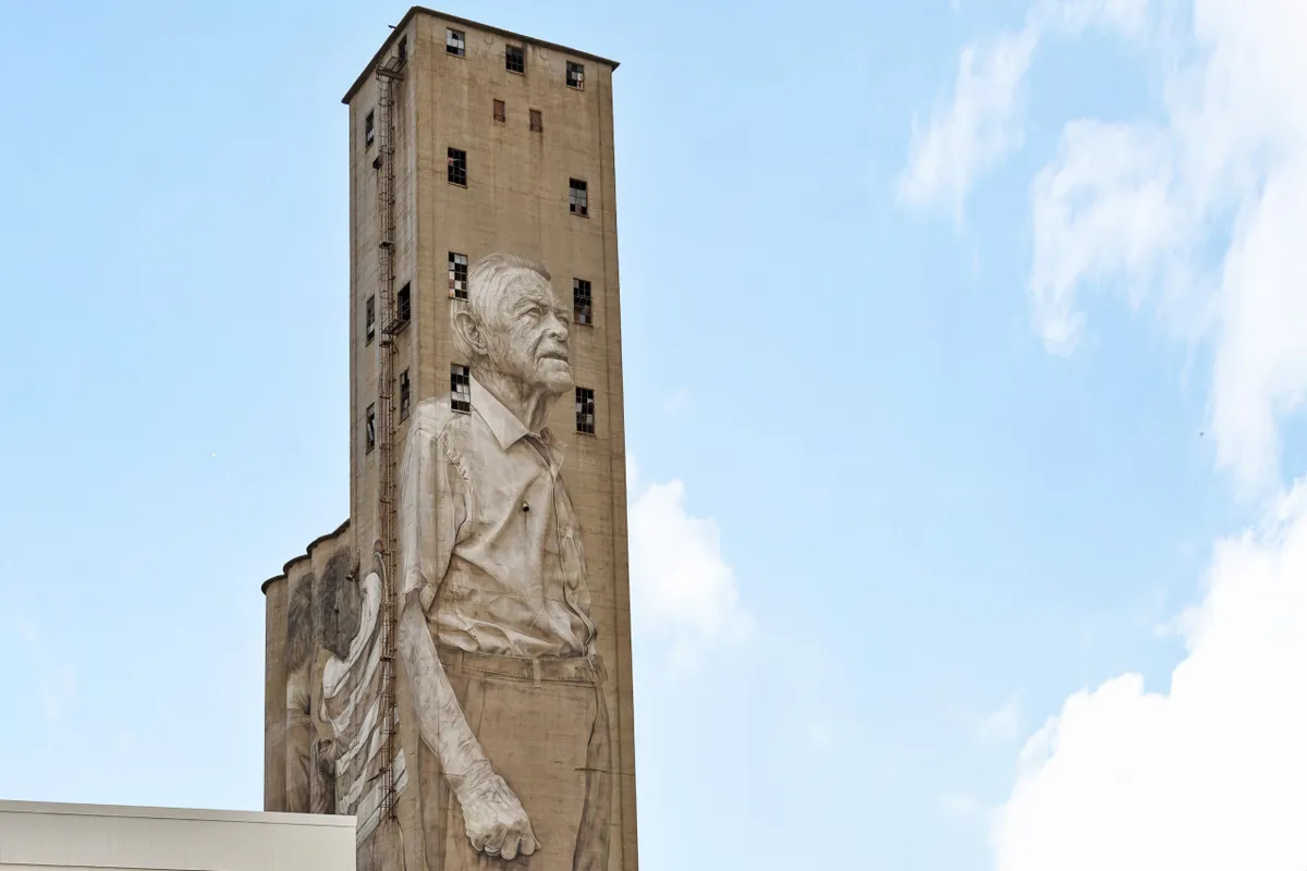 An exterior scene featuring a large mural of an elderly man, with an industrial tower structure nearby. The setting is urban, evoking the creative and energetic vibe of the neighborhood. Passersby might be seen taking photographs or pausing to admire the art, suggesting a lively and walkable community.